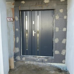 FHU TOMEX Tomasz Słoniewski - Montaż czarnych drzwi zewnętrznych z pionowymi, metalowymi wstawkami w surowym otworze wejściowym, widoczne ocieplenie i resztki zaprawy.