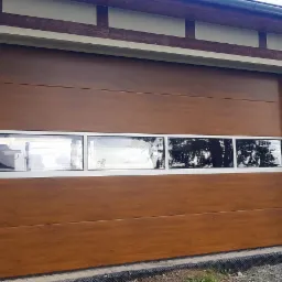 Brązowa brama garażowa segmentowa z poziomymi panelami imitującymi drewno i trzema prostokątnymi oknami z białymi ramami, zamontowana w elewacji budynku z widocznymi elementami drewnianymi.