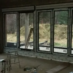 Wnętrze w trakcie montażu okien z czarnymi ramami, widoczne podpory budowlane, drabina i narzędzia, surowy stan wykończenia.