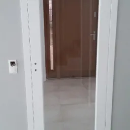 Białe drzwi wewnętrzne z dużym przeszkleniem, widoczna instalacja elektryczna obok ościeżnicy, odbicie w szybie ukazuje kolejne drzwi.