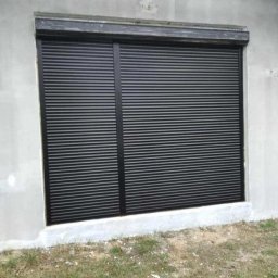 FHU TOMEX Tomasz Słoniewski - Zamknięta, czarna roleta zewnętrzna z poziomych listew zamontowana w oknie budynku o surowym, szarym wykończeniu elewacji. Widoczne fragmenty trawy i ziemi u podstawy ściany.