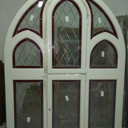FHU TOMEX Tomasz Słoniewski - Białe okno o gotyckim łukowym kształcie z czerwonymi detalami i romboidalnymi szybami, zabezpieczone folią ochronną, widoczne ślady magazynowania.