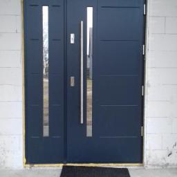 FHU TOMEX Tomasz Słoniewski - Nowoczesne, dwuskrzydłowe, ciemne drzwi wejściowe z przeszkleniami i długimi, pionowymi uchwytami, zamontowane w surowym murze z bloczków betonowych, z napisem 'K+M+B=2017' nad ościeżnicą i czarną...