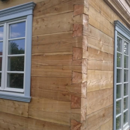 FHU TOMEX Tomasz Słoniewski - Elewacja domu z bali drewnianych z zamontowanymi oknami z białymi ramami i szarymi listwami ozdobnymi, widok z zewnątrz na narożnik budynku i fragment ogrodu.