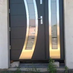 Nowoczesne, grafitowe drzwi zewnętrzne z przeszkleniami o nieregularnym kształcie, obramowane srebrną listwą, osadzone w surowym, betonowym otworze.