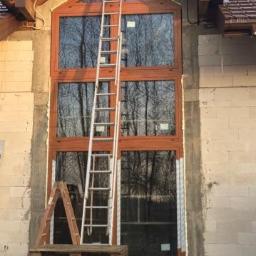 FHU TOMEX Tomasz Słoniewski - Wysokie okno z drewnianą ramą, podzielone na trzy sekcje, z widocznym odbiciem drzew, na tle surowej ściany budynku w trakcie budowy, z drabiną opartą o okno i paletami na dole.