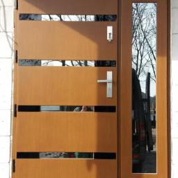 FHU TOMEX Tomasz Słoniewski - Brązowe drzwi zewnętrzne z przeszkleniami i wąskim oknem bocznym, widoczny fragment samochodu i ceglany mur.