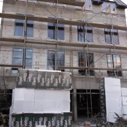 Budynek w trakcie montażu stolarki okiennej, widoczne rusztowania, okna wstawione w surowy mur, na dole składowane paczki styropianu z nadrukiem producenta.