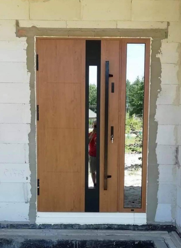 Nowoczesne drzwi zewnętrzne z drewnianym wykończeniem, czarnym akcentem i długim uchwytem, zamontowane w surowym otoczeniu budowlanym w Makowie Mazowieckim, z odbiciem fotografa w szybie.