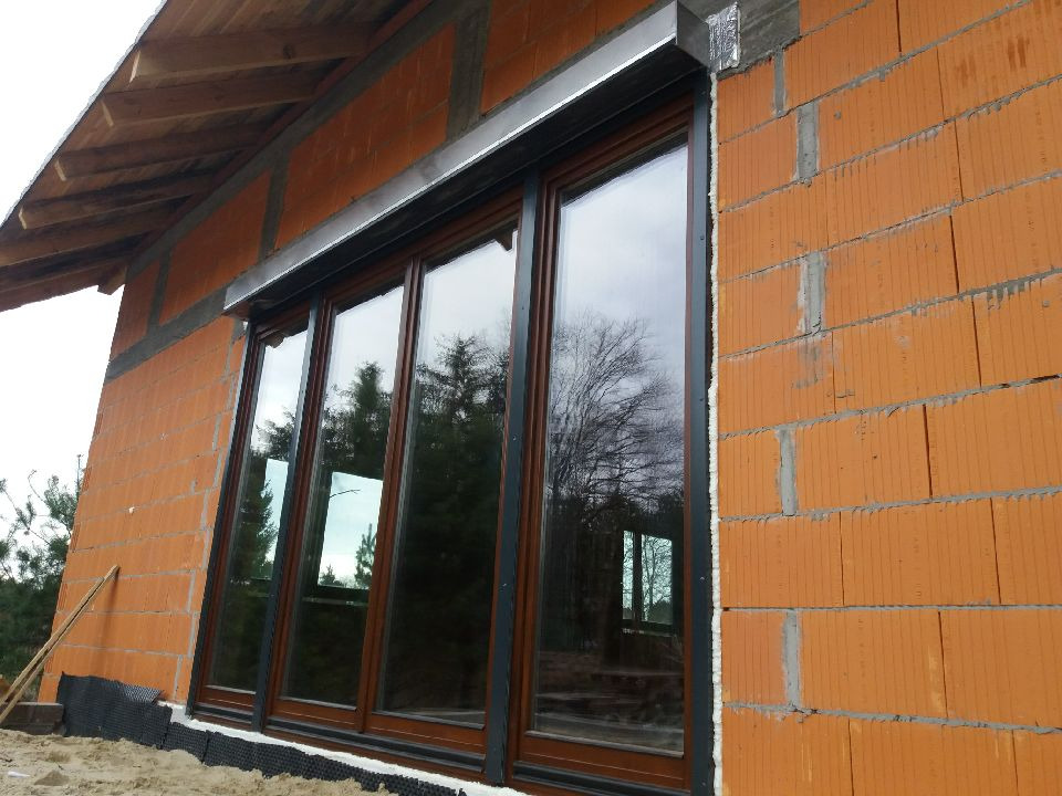 Czterodzielne okno z brązową ramą i szarą roletą zewnętrzną zamontowane w surowej, ceglanej ścianie budynku w trakcie budowy.