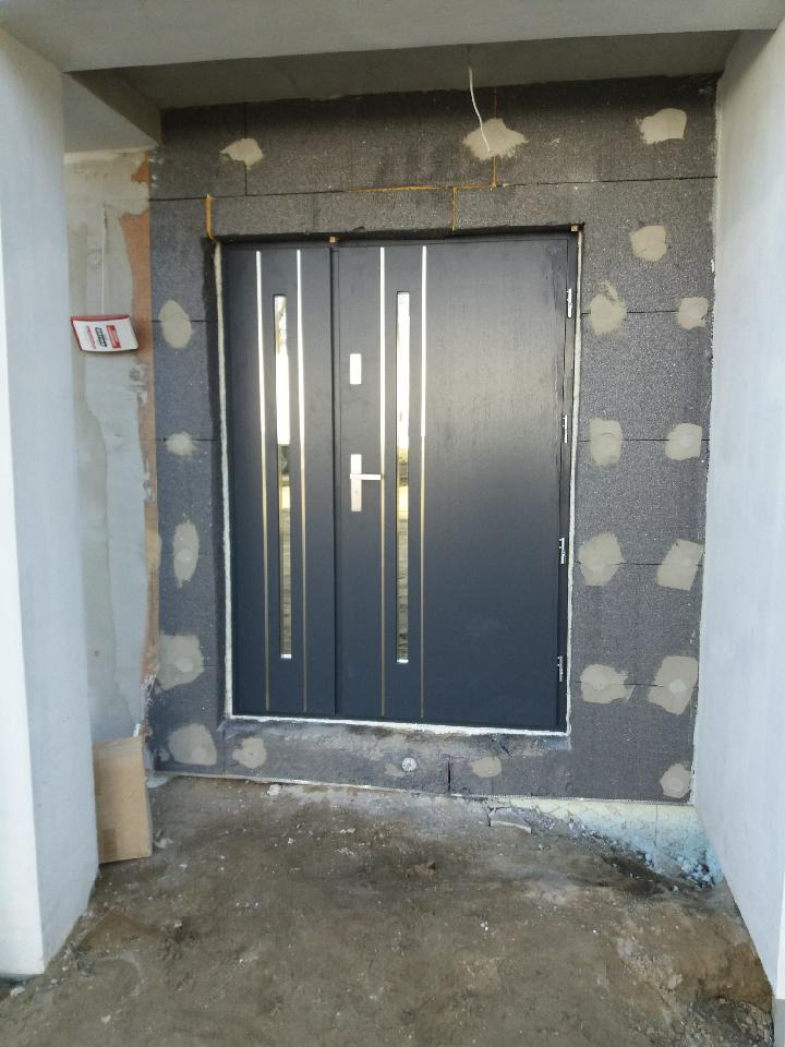 Montaż czarnych drzwi zewnętrznych z pionowymi, metalowymi wstawkami w surowym otworze wejściowym, widoczne ocieplenie i resztki zaprawy.