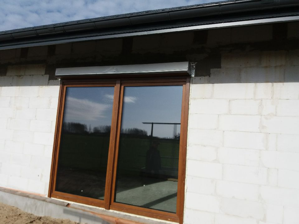 Brązowe okno przesuwne z roletą zewnętrzną w surowym stanie, widoczne ślady po montażu na ścianie z białego bloczka.