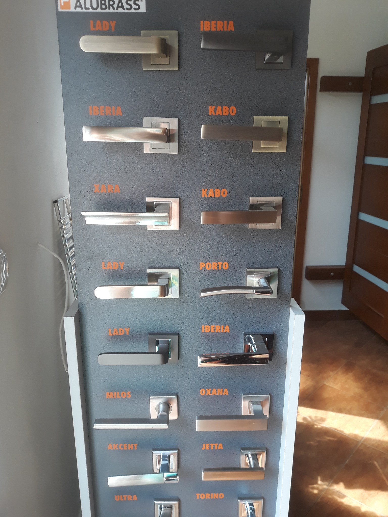 Ekspozycja różnych modeli klamek do drzwi wewnętrznych, prezentowanych na szarym panelu z nazwami serii: Lady, Iberia, Kabo, Xara, Porto, Milos, Oxana, Akcent, Jetta, Ultra, Torino