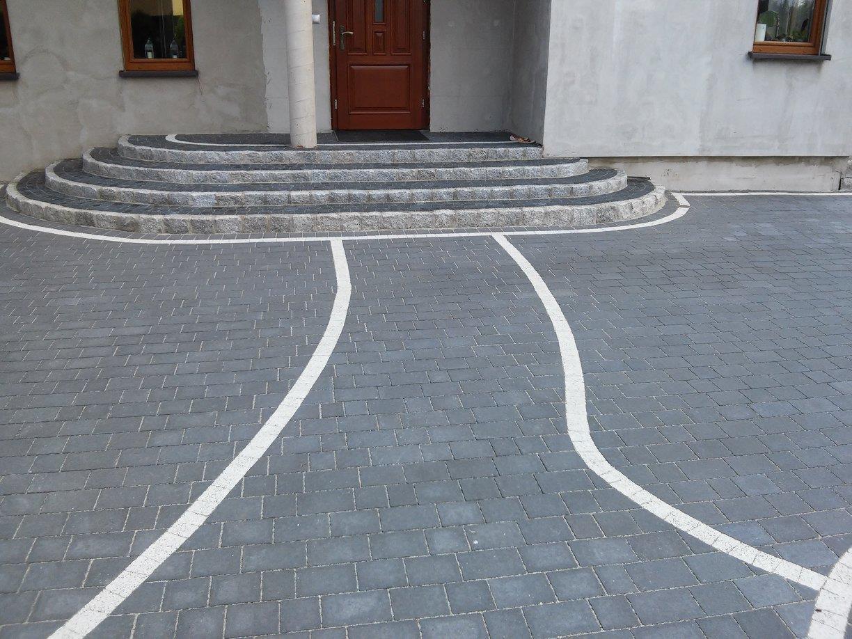 Ułożona szara kostka brukowa z białymi liniami wyznaczającymi ścieżkę w Godowie, prowadząca do schodów wejściowych z ciemnymi stopniami i jasnymi obrzeżami.