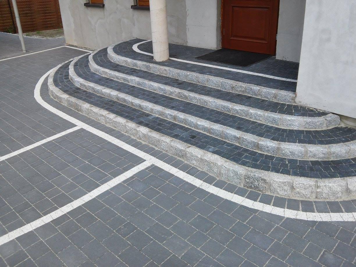 Schody wejściowe wyłożone ciemną kostką granitową z jasnymi obrzeżami, prowadzące do drzwi wejściowych budynku z szarą elewacją i kolumną.