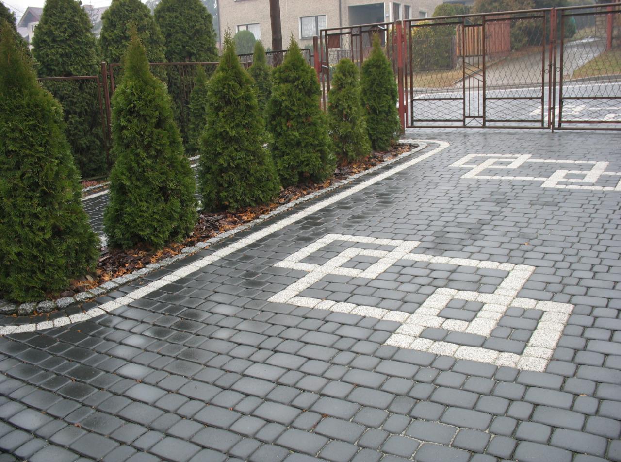 Ułożona kostka brukowa w kolorze grafitowym z białym wzorem geometrycznym, obrzeże z szarego kamienia, rząd tuj wzdłuż ogrodzenia.