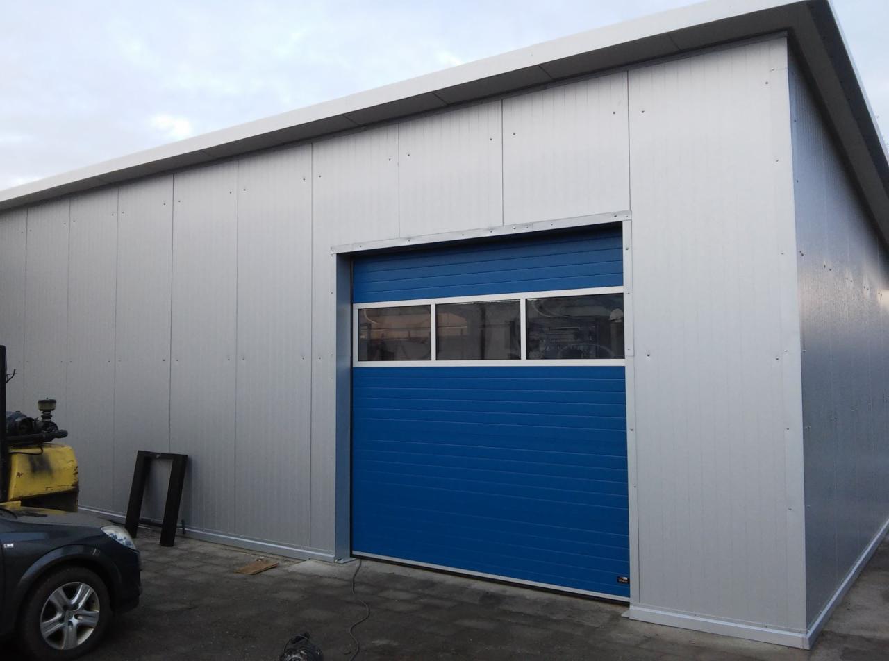 Nowoczesny budynek przemysłowy z szarej blachy trapezowej, z niebieską bramą segmentową z trzema oknami, widok z zewnątrz.