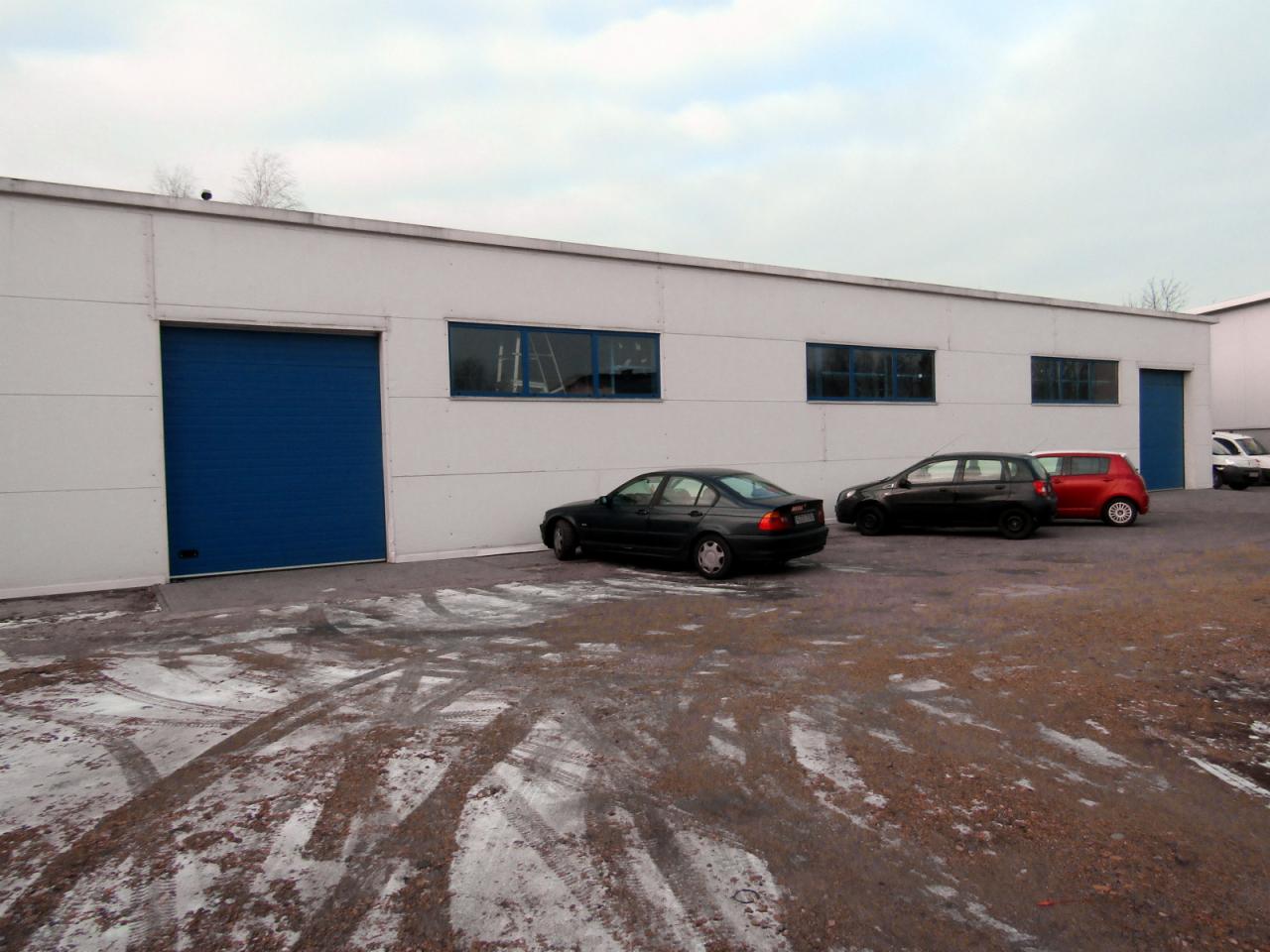 Biały budynek przemysłowy z niebieską bramą segmentową, kilkoma oknami i trzema zaparkowanymi samochodami przed nim, częściowo pokryty śniegiem teren.