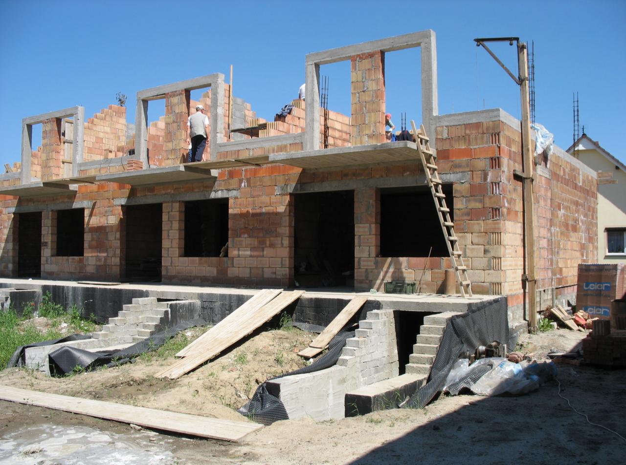 Budowa domu jednorodzinnego z czerwonej cegły, widoczne otwarte okna, schody wejściowe i rusztowania, dwóch pracowników na górze, błękitne niebo w tle.