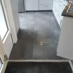 Szare, matowe płytki podłogowe w kuchni, widoczne z perspektywy wejścia, z fragmentem białych szafek kuchennych i srebrnej lodówki w tle.