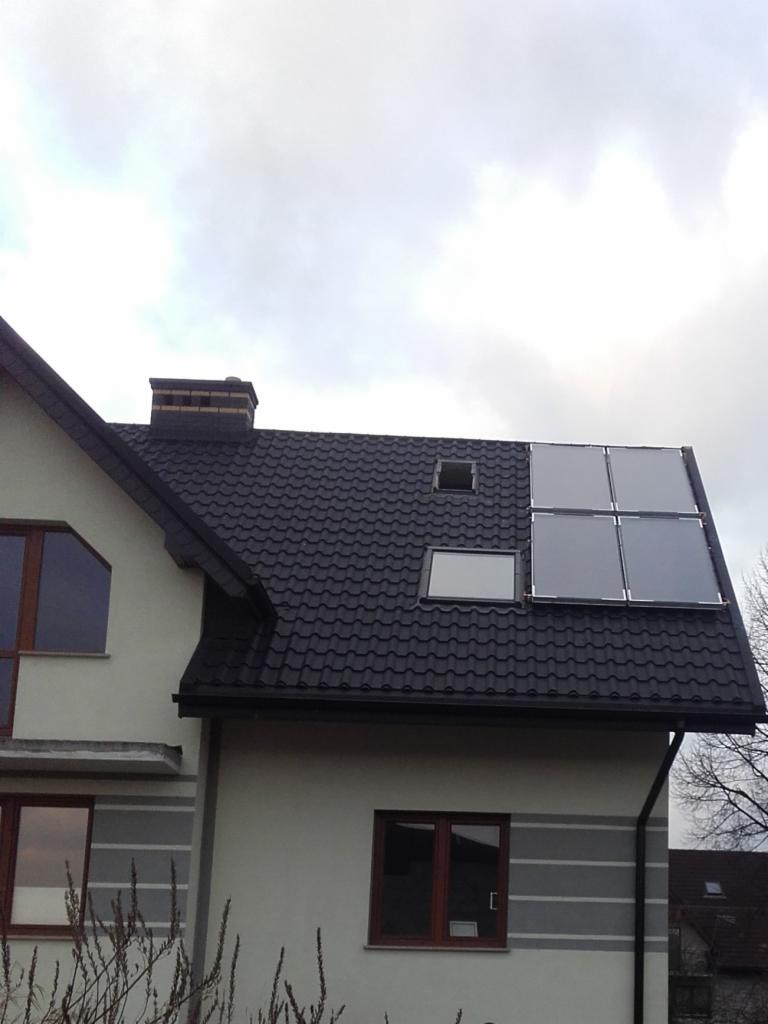 Dom jednorodzinny z ciemną dachówką, oknem dachowym i panelami słonecznymi na dachu, widoczne z poziomu gruntu w pochmurny dzień.