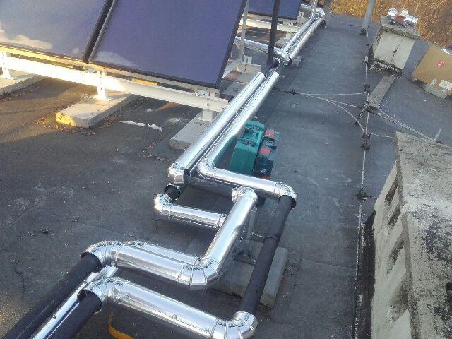 Instalacja solarna na dachu: widoczne panele słoneczne, rury w srebrnej izolacji, zielona pompa obiegowa, elementy mocujące i komin wentylacyjny w tle.