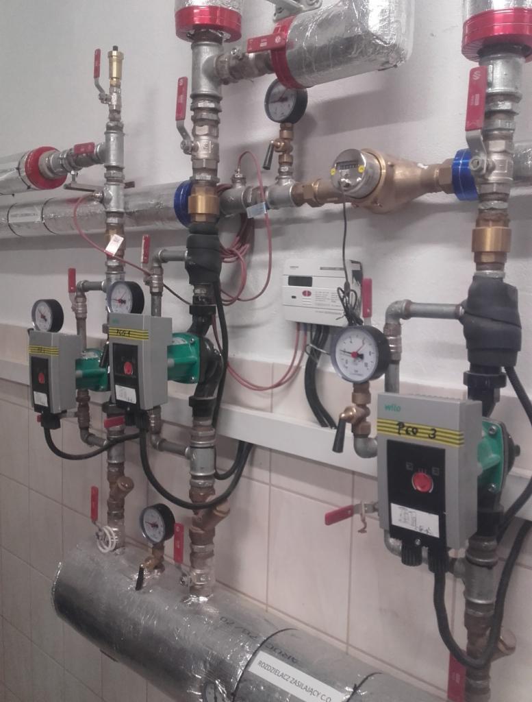 Rozbudowana instalacja centralnego ogrzewania z widocznymi rurami, zaworami, manometrami, pompami obiegowymi Wilo i licznikiem ciepła zamontowana na ścianie wyłożonej płytkami.