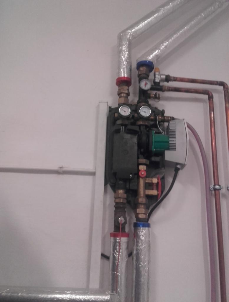 Węzeł cieplny z pompą obiegową i manometrami, rury miedziane i izolowane, instalacja w pomieszczeniu technicznym.