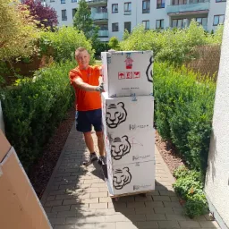 Uśmiechnięty mężczyzna w pomarańczowej koszulce i rękawicach transportuje na w&oacute;zku stos kartonowych pudeł z logo Asian Tigers Mobility na tle nowoczesnego apartamentowca i zieleni.