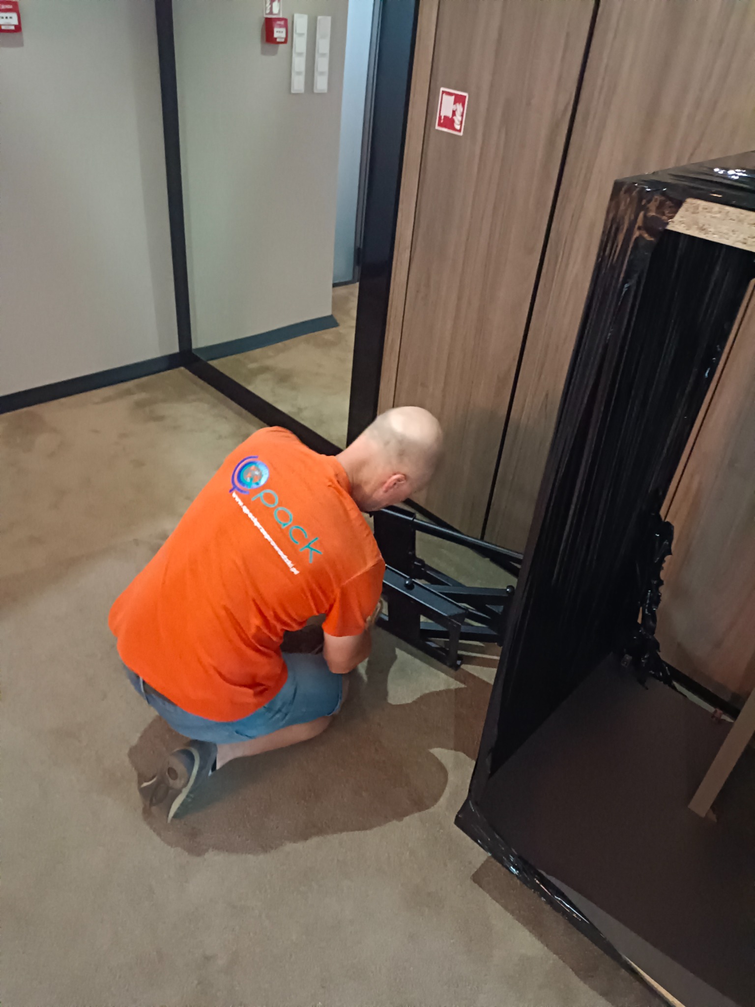 Pracownik w pomarańczowej koszulce z logo firmy transportowej klęczy przy zabezpieczonym folią meblu, prawdopodobnie podczas przeprowadzki wewnątrz budynku.