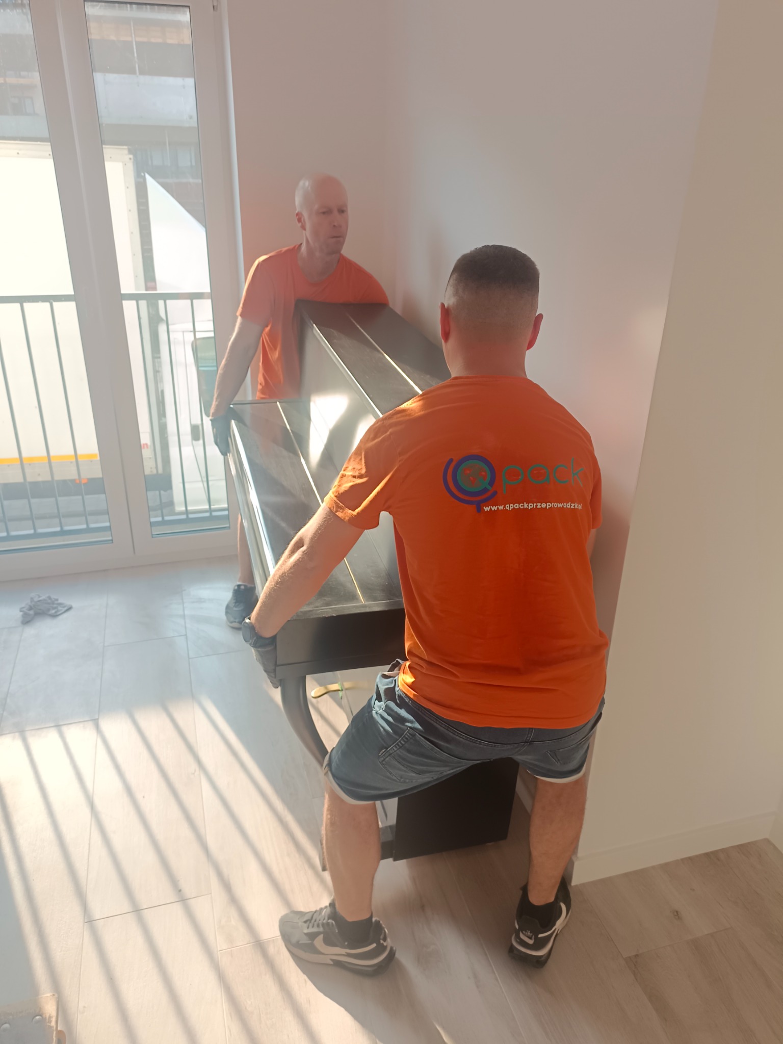 Dwóch mężczyzn w pomarańczowych koszulkach z logo firmy przeprowadzkowej, w rękawicach, wnosi czarny fortepian do jasnego pomieszczenia z dużymi oknami, przez które widać zaparkowaną ciężarówkę.