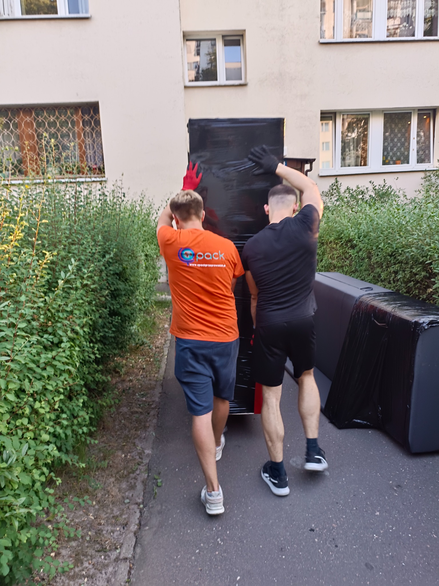 Dwóch mężczyzn w roboczych ubraniach, z których jeden ma pomarańczową koszulkę z logo firmy, a drugi czarną koszulkę, przenosi zapakowane w czarną folię meble na zewnątrz budynku.