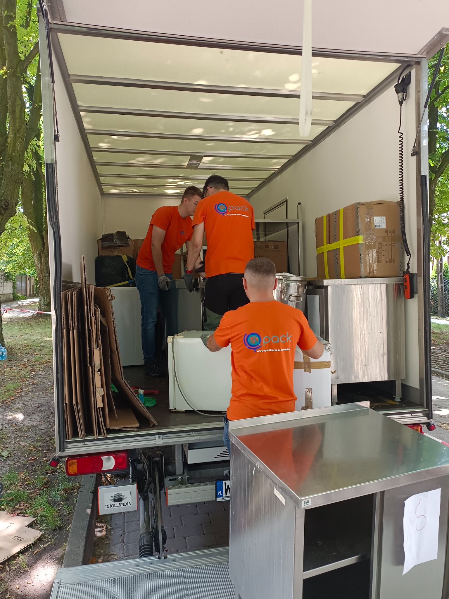 Trzech mężczyzn w pomarańczowych koszulkach z logo firmy przeprowadzkowej ładuje białe sprzęty AGD i kartony do wnętrza dużej furgonetki z otwartą rampą załadunkową na zewnątrz.