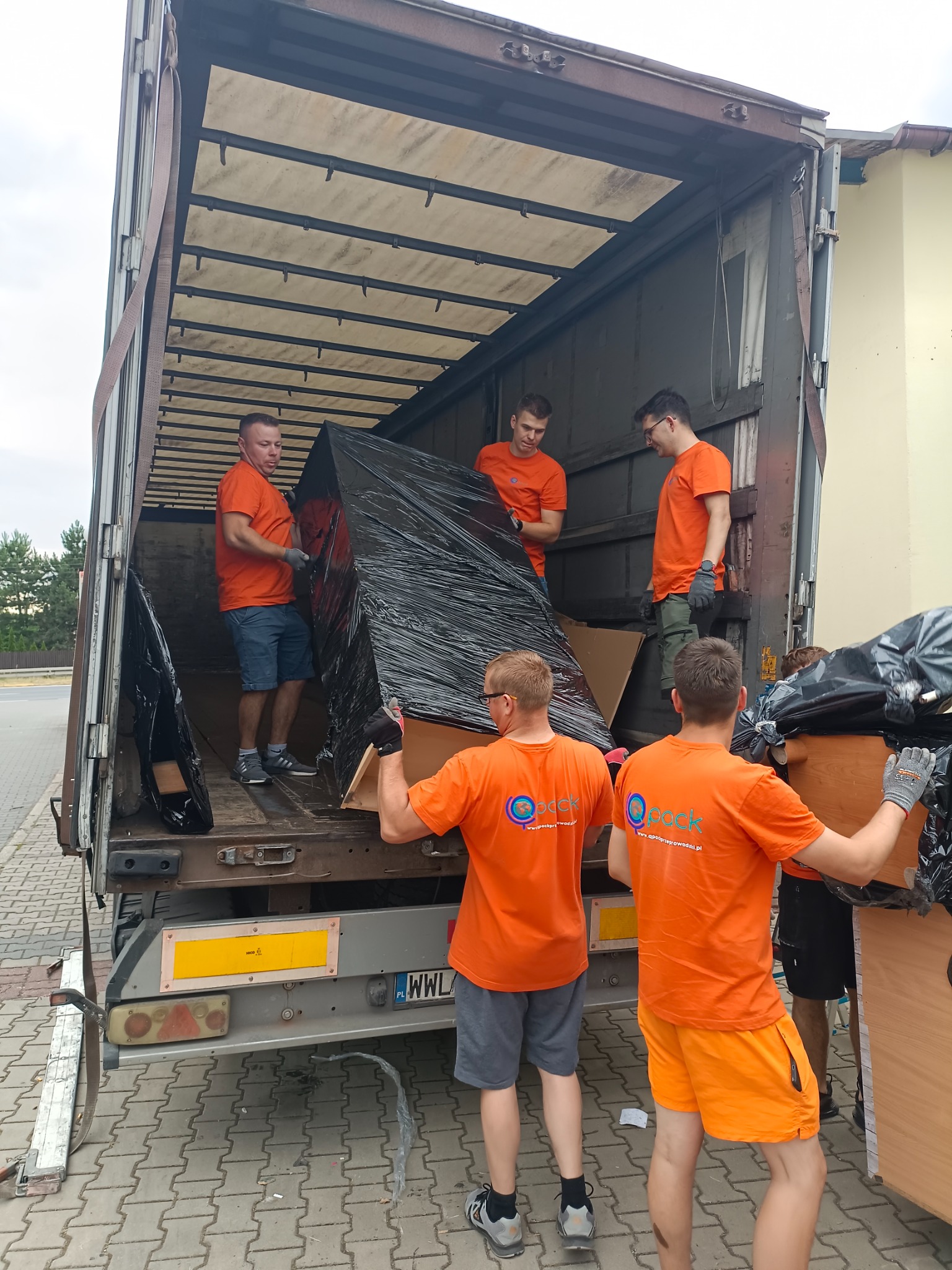 Czterech mężczyzn w pomarańczowych koszulkach z logo firmy transportowej ładuje meble owinięte czarną folią do wnętrza ciężarówki z otwartą przestrzenią ładunkową.
