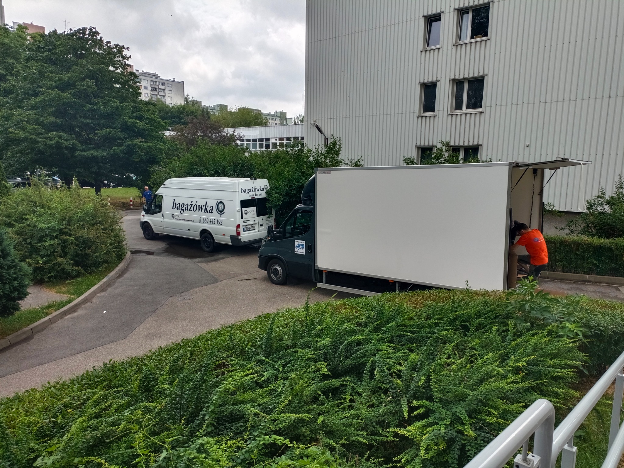 Dwa busy transportowe zaparkowane obok budynku mieszkalnego, jeden z otwartą przestrzenią ładunkową i pracownikiem w pomarańczowej koszulce w środku, drugi z logo firmy 'bagażówka' na boku.