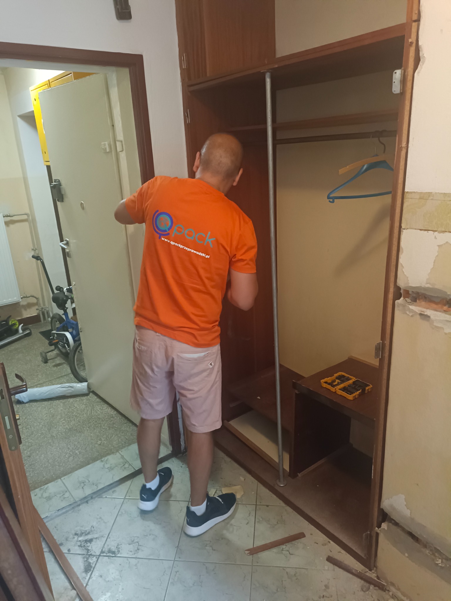 Pracownik firmy przeprowadzkowej w pomarańczowej koszulce z logo, demontuje drewnianą szafę w trakcie przeprowadzki, widoczne narzędzia, fragment roweru w tle.