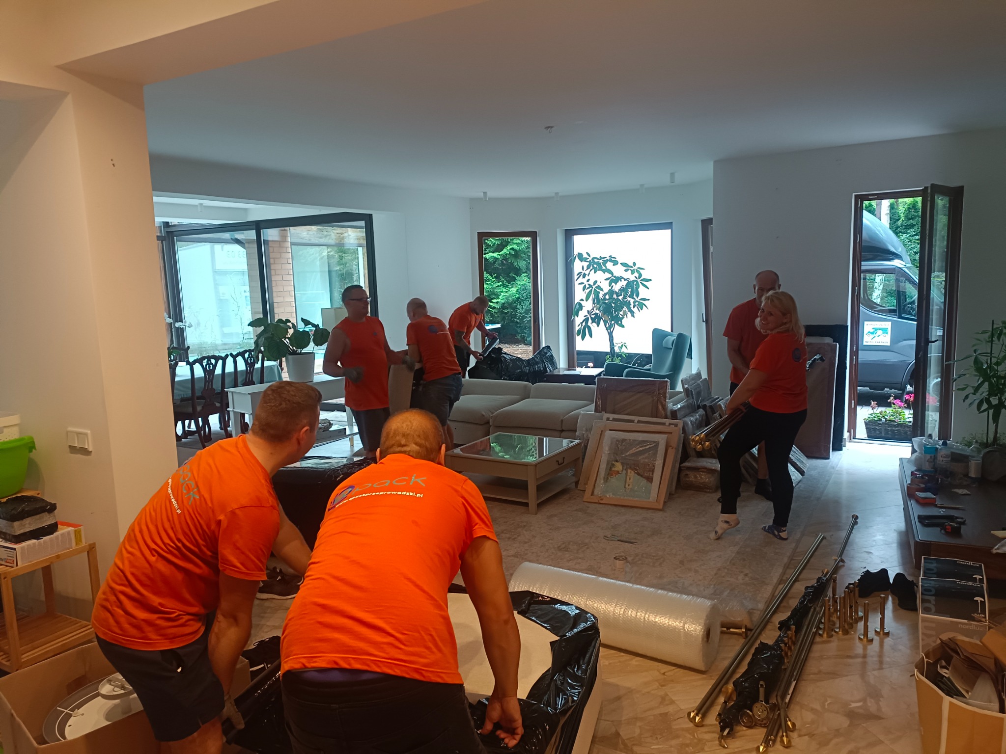 Ekipa przeprowadzkowa w pomarańczowych koszulkach pakuje meble w przestronnym salonie z dużymi oknami i widokiem na ogród, w tle zaparkowany bus.