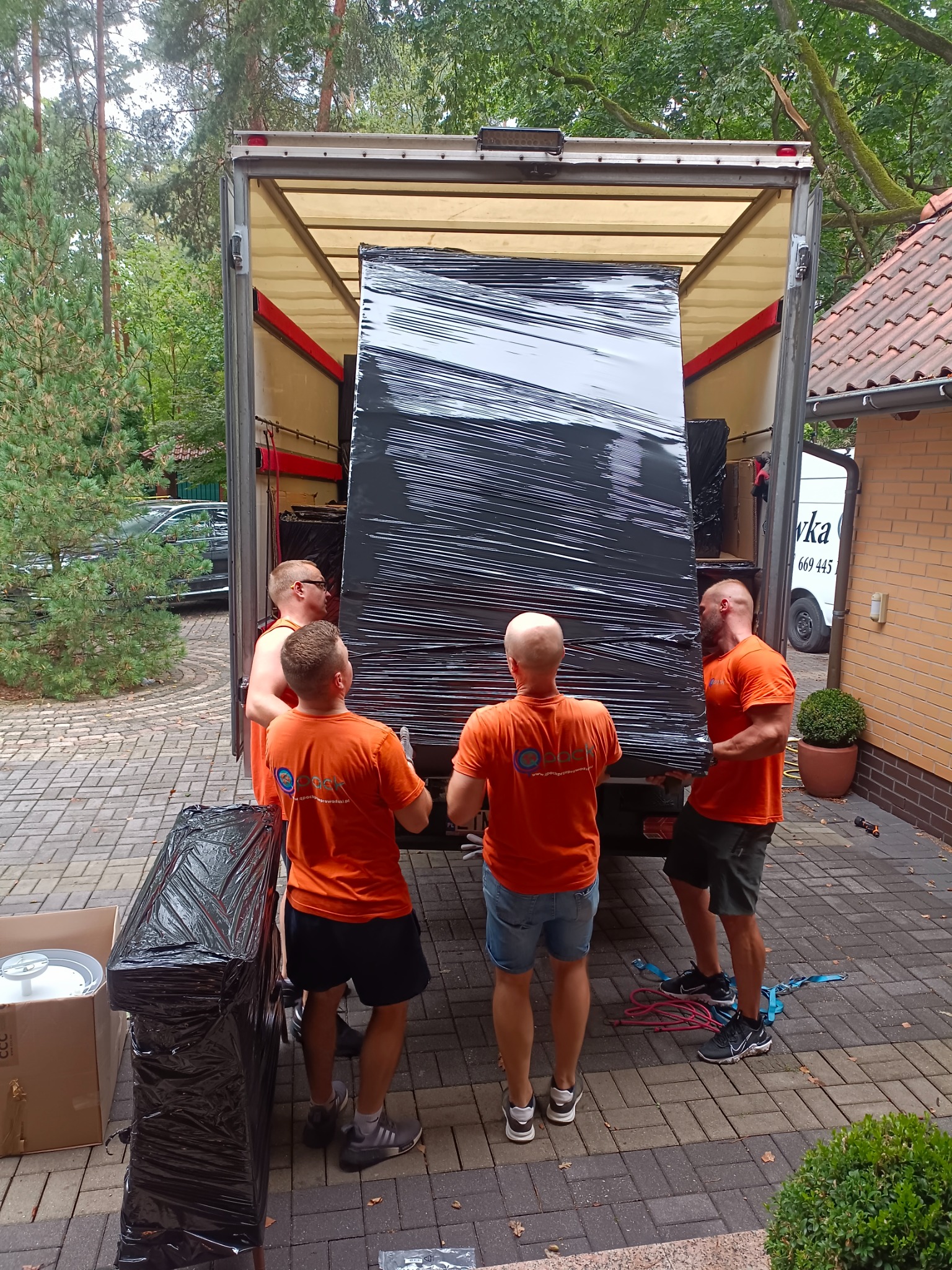 Czterech mężczyzn w pomarańczowych koszulkach wnosi do furgonetki owinięty czarną folią mebel na tle domu z cegły i zieleni.