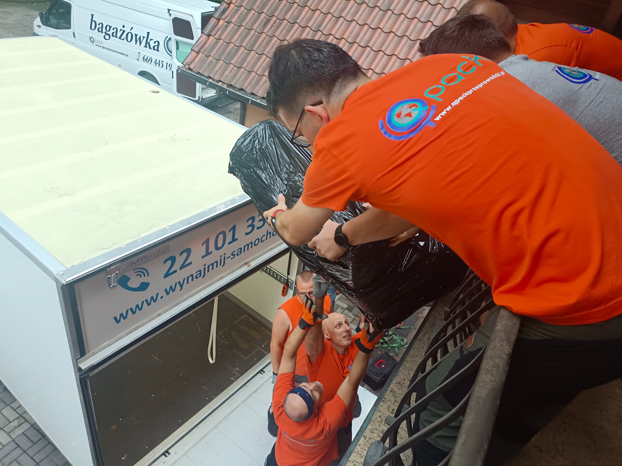 Ekipa czterech mężczyzn w pomarańczowych koszulkach z logo firmy transportowej, przenosi zabezpieczony folią mebel z balkonu do wnętrza samochodu dostawczego z napisem 'wynajmij-samochod', widok...