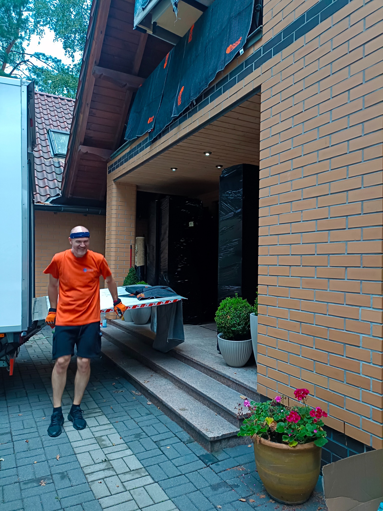 Pracownik w pomarańczowej koszulce i rękawicach, z uśmiechem, stoi obok ciężarówki przed domem z zapakowanymi meblami, przygotowanymi do transportu.