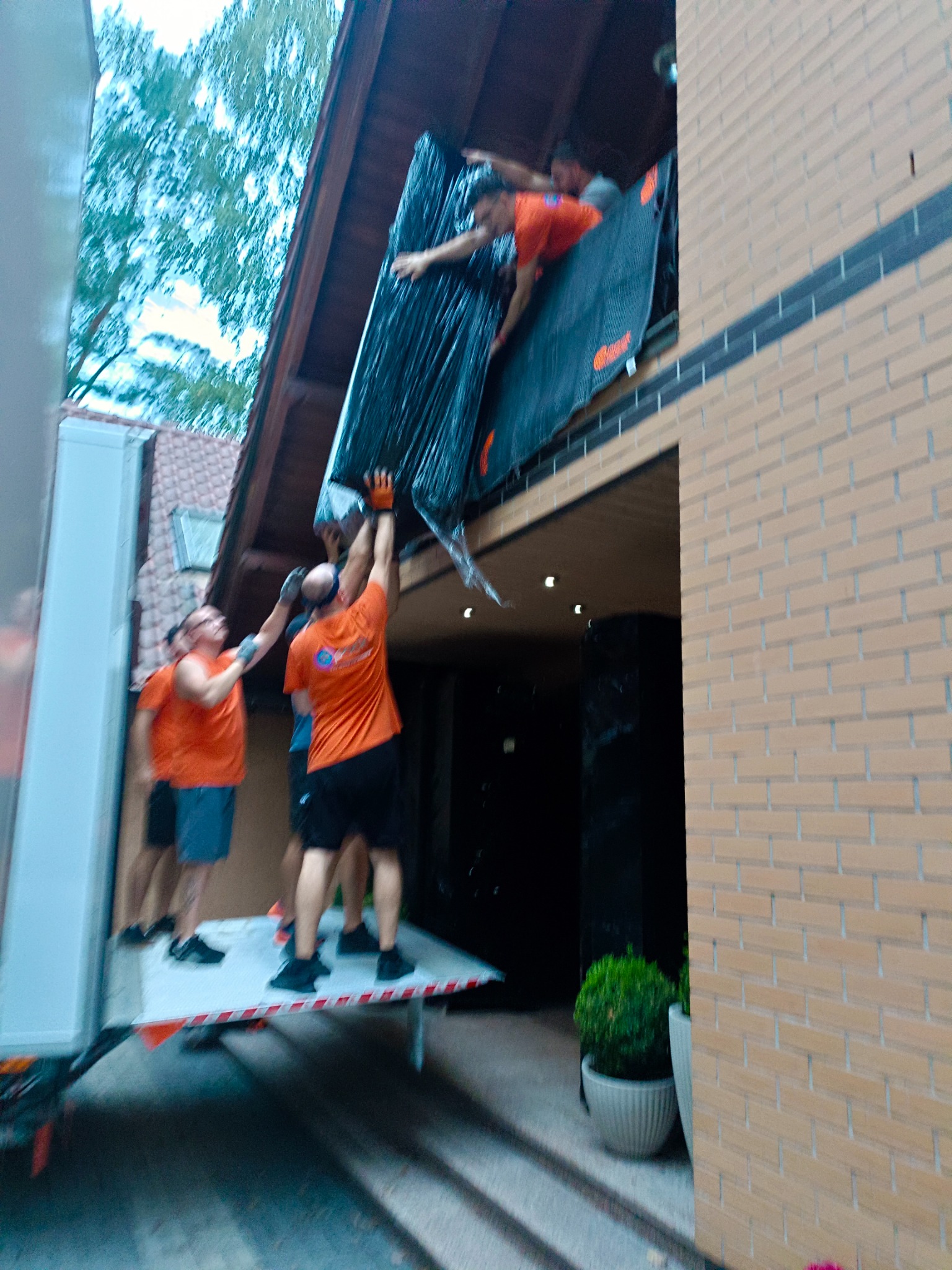 Ekipa w pomarańczowych koszulkach przenosi zabezpieczony mebel z platformy ciężarówki do budynku z cegły, widok z dołu, z widocznymi drzewami w tle.