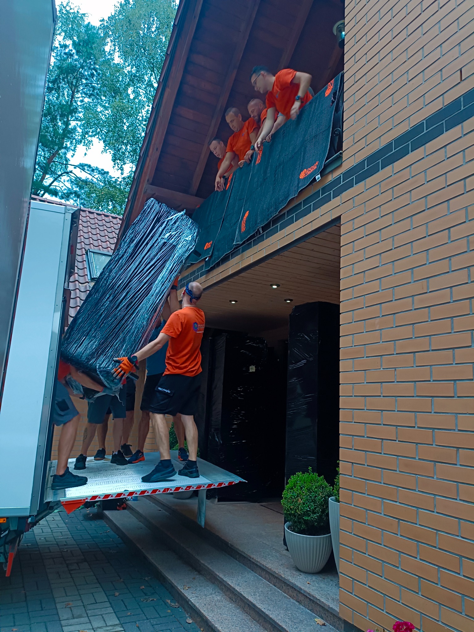 Grupa mężczyzn w pomarańczowych koszulkach przenosi mebel owinięty folią z balkonu domu do windy samochodu dostawczego, widoczny fragment elewacji z cegły i drzewa w tle.