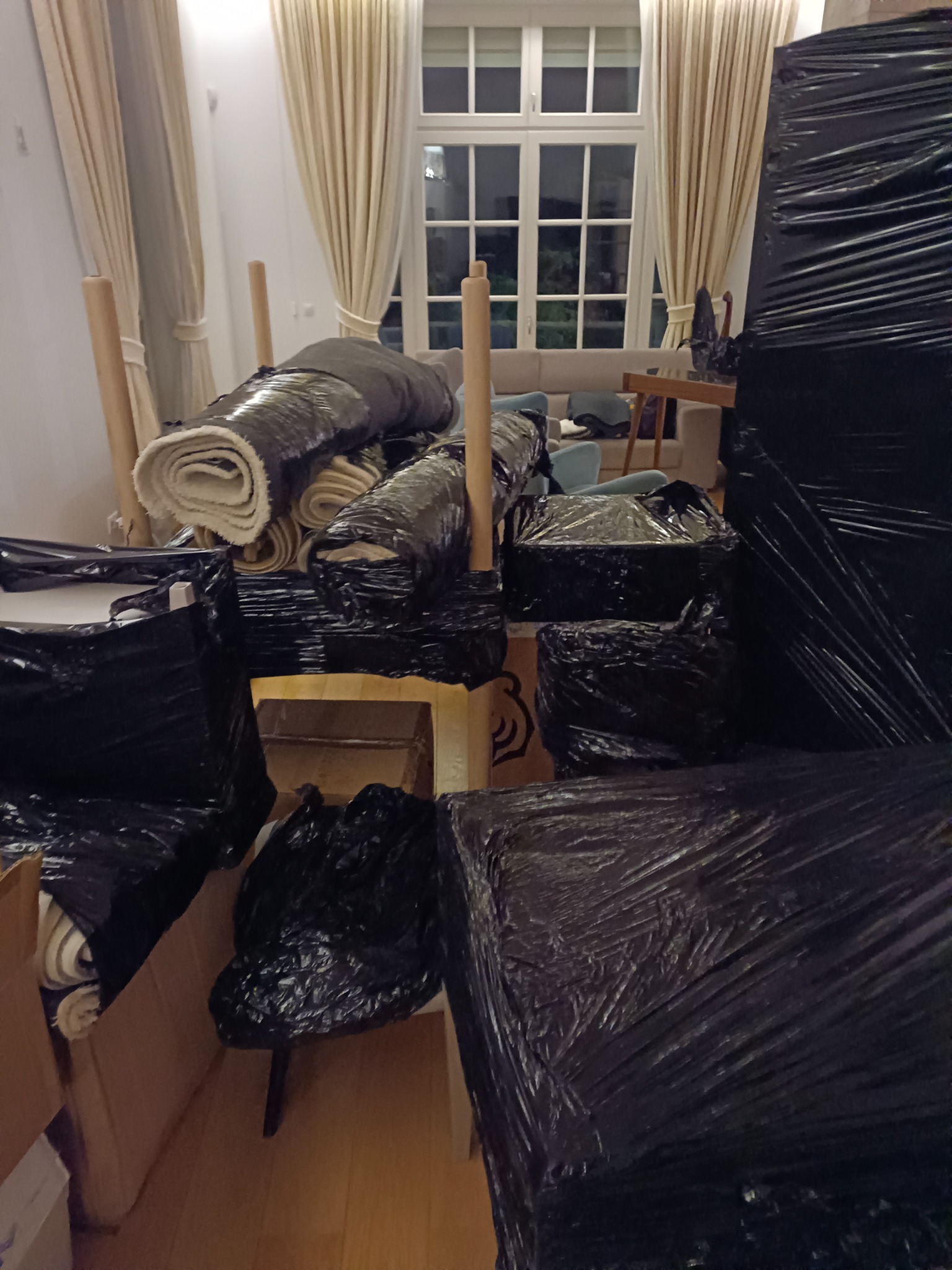 Pokój w trakcie pakowania przed przeprowadzką: meble i zwinięte dywany zabezpieczone folią i umieszczone w kartonach, widoczne jasne zasłony i okno.