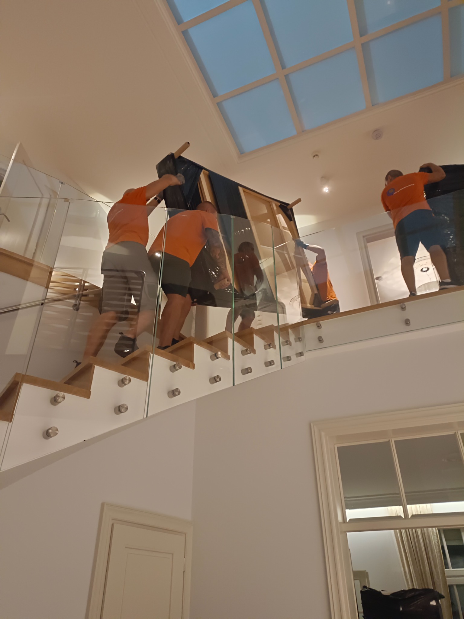 Czterech mężczyzn w pomarańczowych koszulkach wnosi mebel zabezpieczony folią po schodach z szklaną balustradą w jasnym wnętrzu z oknem dachowym.
