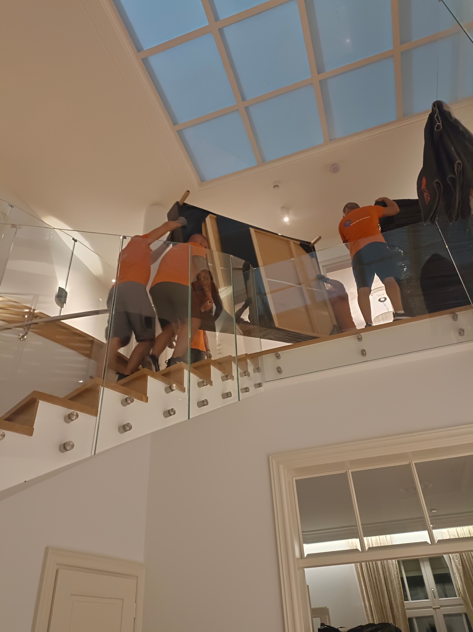 Ekipa w pomarańczowych koszulkach wnosi obraz po schodach z drewnianymi stopniami i szklaną balustradą w jasnym wnętrzu z oknem dachowym.