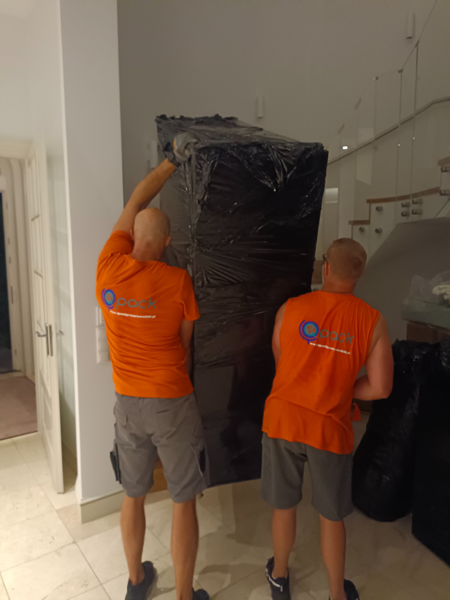 Dwóch pracowników w pomarańczowych koszulkach z logo firmy transportowej niesie zabezpieczony folią mebel w jasnym wnętrzu domu z marmurową podłogą i schodami ze szklaną balustradą.