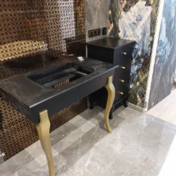 Czarna umywalka nablatowa z wyfrezowanym miejscem na baterię, wsparta na złotych, giętych nogach, obok czarna komoda z szufladami na tle wzorzystych płytek ściennych.