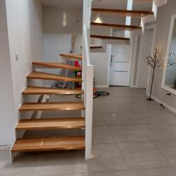 Usługi Stolarskie Marcin Biesiadecki - Nowoczesne schody z drewnianymi stopniami i białymi wspornikami w jasnym wnętrzu z oknem dachowym, widoczne dekoracje i odkurzacz w tle.