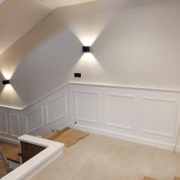 Usługi Stolarskie Marcin Biesiadecki - Wnętrze domu w trakcie wykańczania z białą boazerią ścienną z ozdobnymi ramkami, oświetleniem punktowym i fragmentem schodów. Na podłodze leżą kartony ochronne, a w rogu stoi rozłożona drabina.