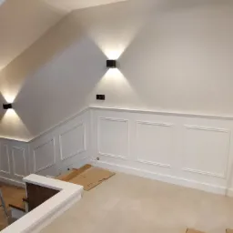 Wnętrze domu w trakcie wykańczania z białą boazerią ścienną z ozdobnymi ramkami, oświetleniem punktowym i fragmentem schodów. Na podłodze leżą kartony ochronne, a w rogu stoi rozłożona drabina.